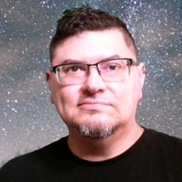 Male with dark hair, glasses and a goatee wears a black shirt and stands in front of a starry background.